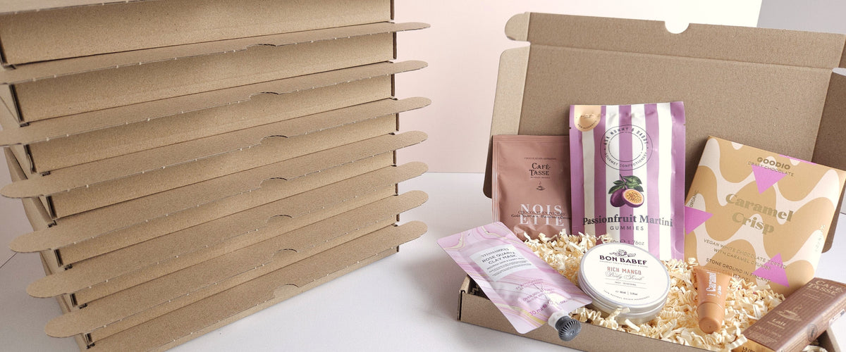 Stapel brievenbusdozen van CaDo Depot naast een geopende cadeaubox met chocolade, gummies en verzorgingsproducten.